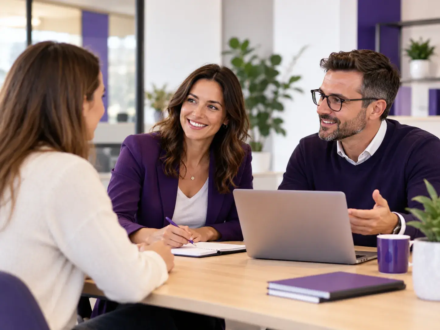 Zwei Mitarbeiter beraten eine Besucherin in einem modernen Büro mit Laptop und violetten Akzenten.